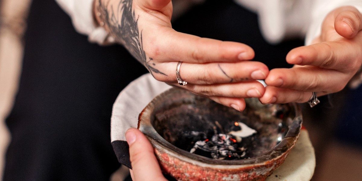 Tattooed hands with light-toned skin cup together over a smudging bowl.