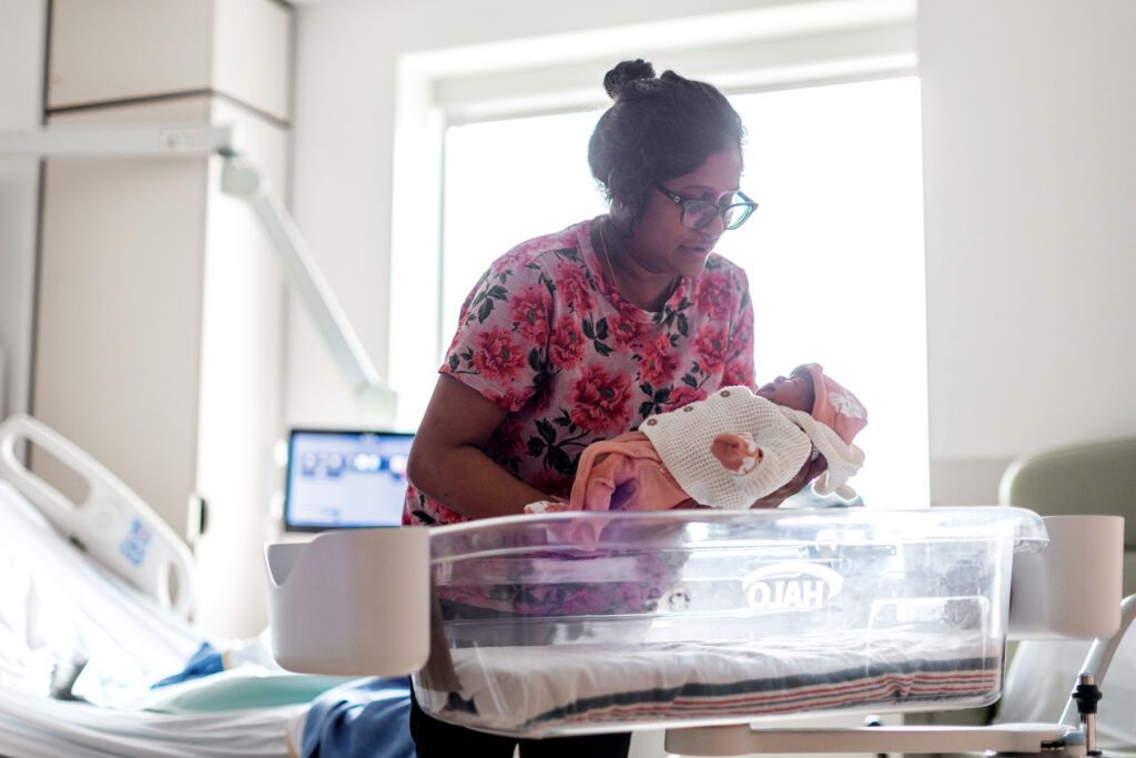 A nurse wears a pink floral shirt. She has dark-toned skin and wears her dark brown hair in a bun. She looks over her dark rimmed glasses while gently placing a newborn baby into a hospital bassinet.