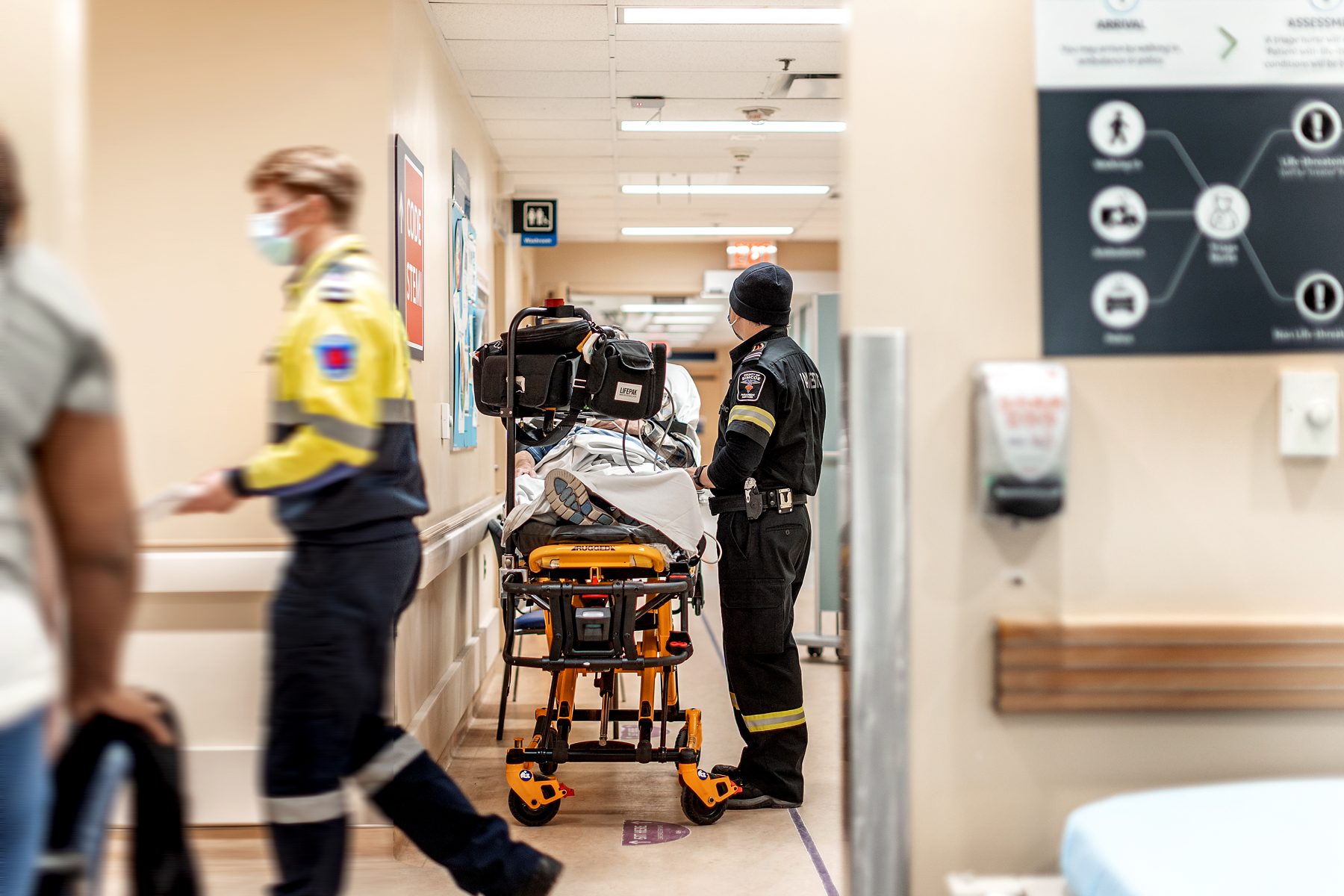 A hospital hallway holds a portable Emergency Services gurney with a patient laying on it. The EMS worker stands next to the gurney and their medical bag hangs from a hook attached to the gurney.