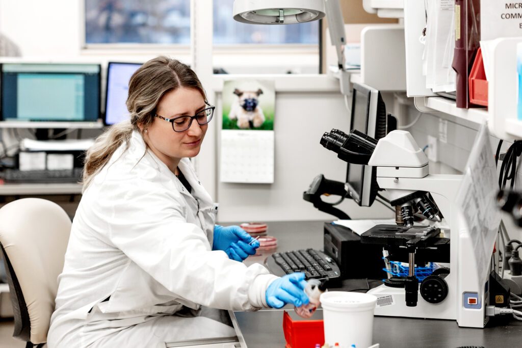 A health care worker wears blue medical gloves and a white lab coat while working at a large microscope. She is holding a piece of the microscope out.