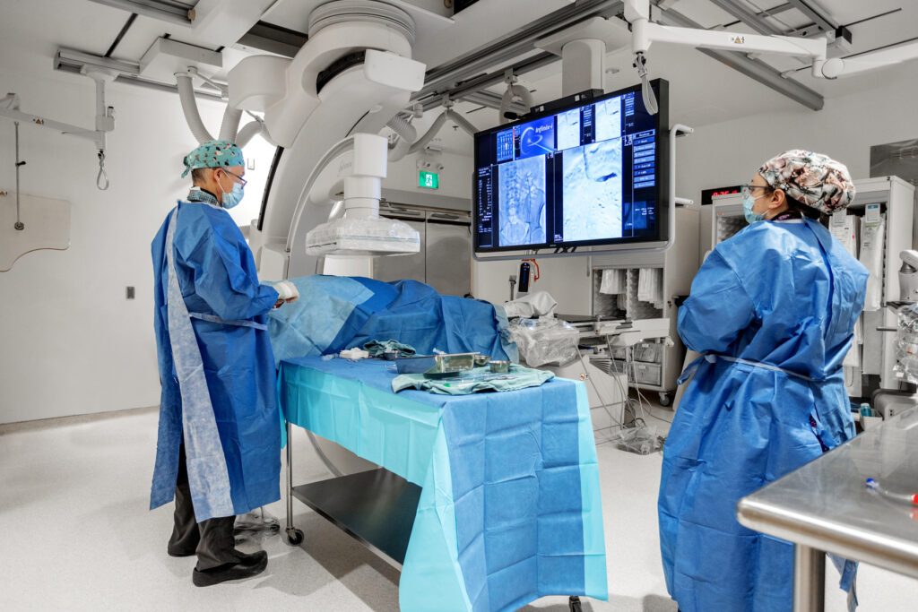 Two health care workers stand in an operating room surrounded by sterile equipment. They look at a large screen with medical images on it.