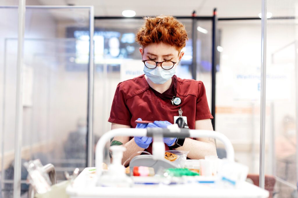 A health care worker holds an item in their hands while standing at a cart with various medical implements on it. The professional wears red scrubs, round glasses, and a face mask. They have short red hair, light toned-skin and each eyebrow is pierced.