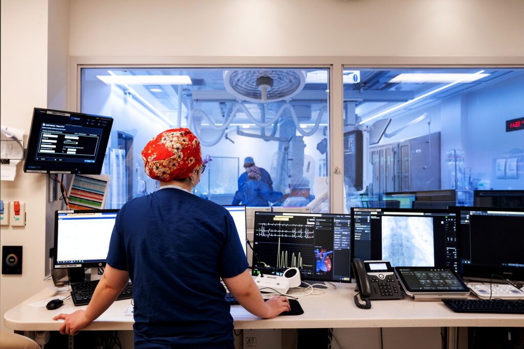 A health care worker stands at a long desk with many screens of information while looking into an operating room at two other medical professionals.