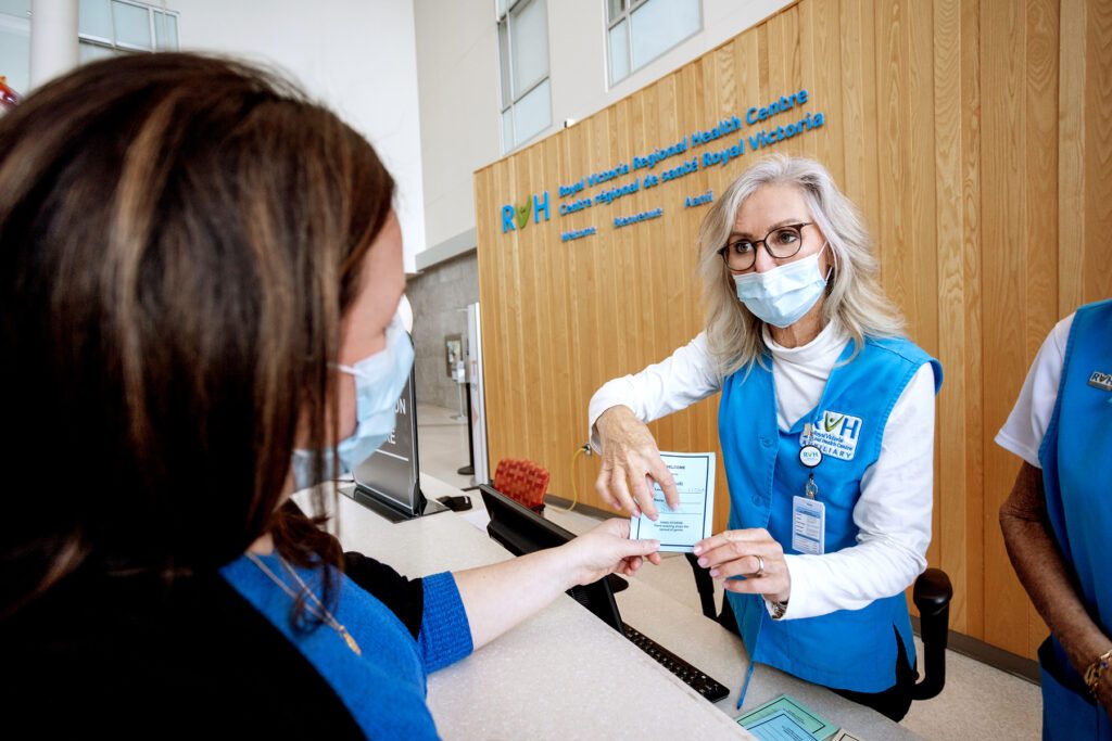 An RVH worker holds out a small piece of paper to a patient and points at the paper. The worker wears a blue vest with RVH branding, glasses, and a medical mask. She has long, light hair and light-toned skin.