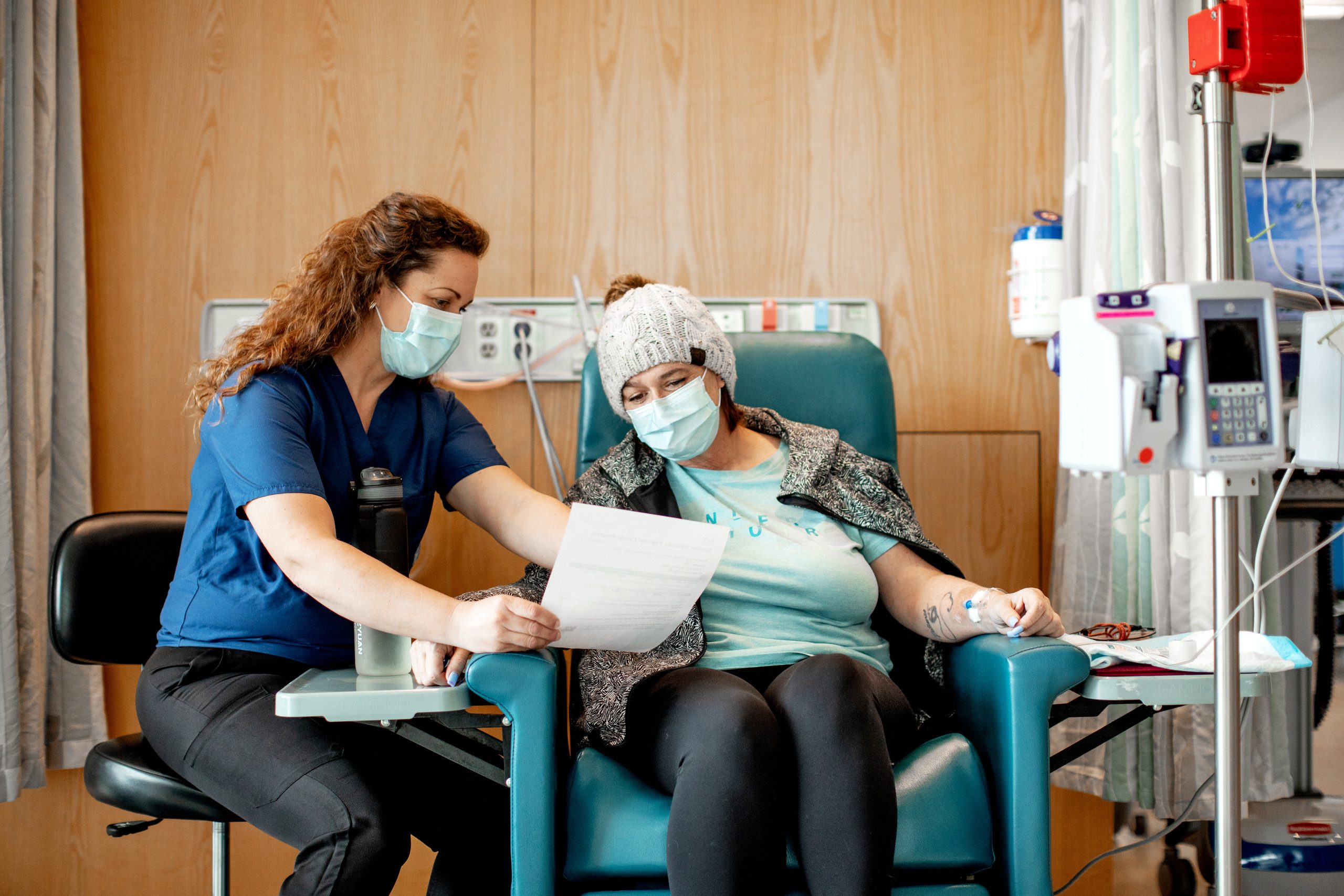 A health care worker with curly brown hair wears a face mask and holds out a sheet of paper to a patient in a chair. The patient wears a knitted hat and has a jacket draped over their shoulders. The patient also wears a medical face mask while sitting next to an IV.