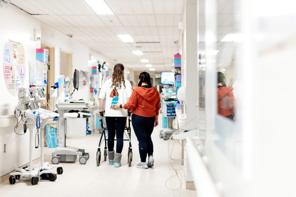 A patient walks down the hospital hallway with a mobility aid while a second person follows with their hand supporting their back.