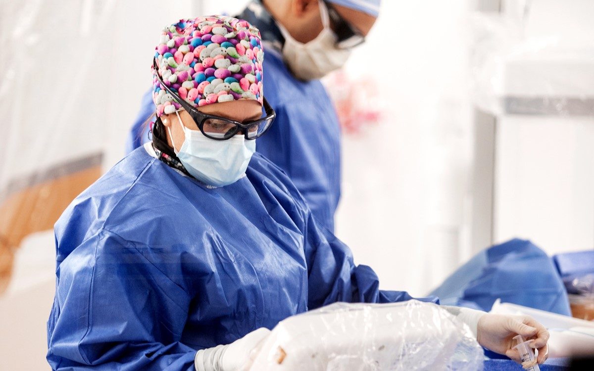 2 health care workers wear blue medical suits and bandanas that cover their heads. They bot wear goggles over their eyes and masks cover their faces as they look down at what they're doing in an area covered with plastic.