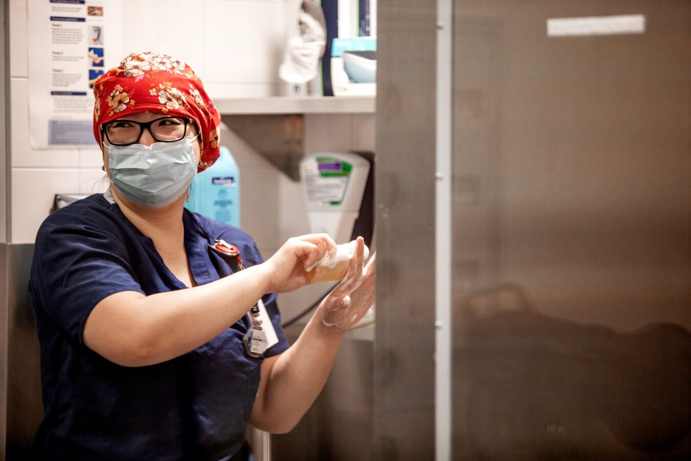 A health care worker stands at a sink, scrubbing their hands with soap. They wear a floral bandanna over their hair, black rimmed glasses, and a face mask. They have light-toned skin and blue scrubs.