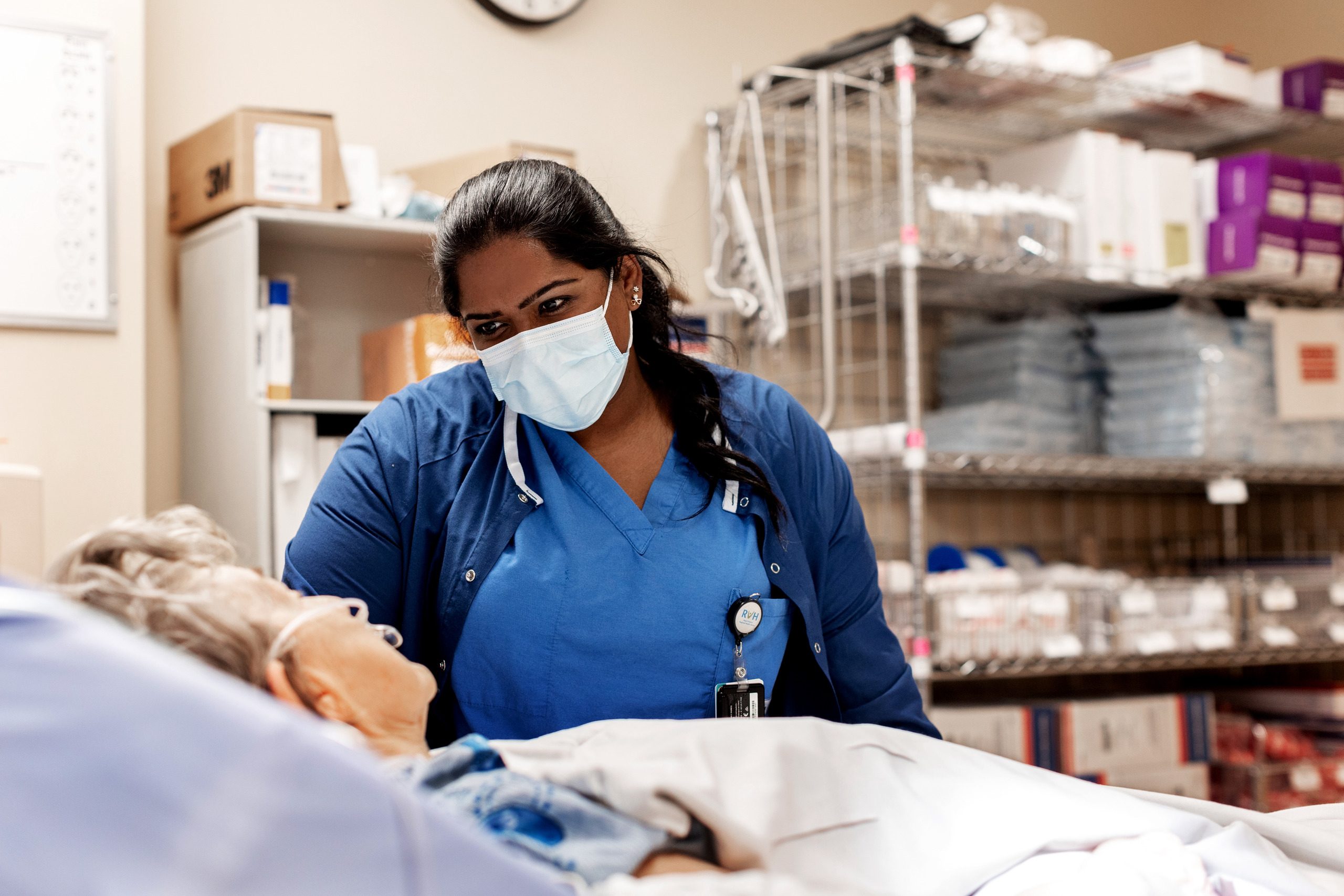 A healthcare worker wearing blue scrubs and a face mask stands next to a patient's bed and looks down at them.