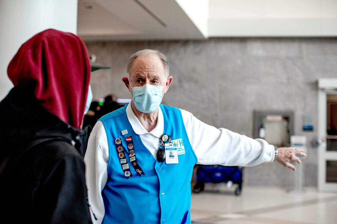 An RVH worker greets a patient and directs them with an outstretched hand. The worker wears a face mask and blue vest with RVH branding. He has short grey hair and light-toned skin.